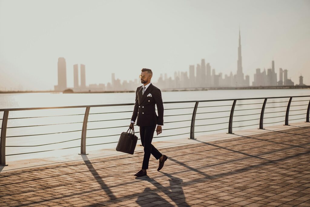 Man in a dark suit walking with a work bag along a waterfront promenade, Burj Khalifa and the Dubai skyline hazy behind