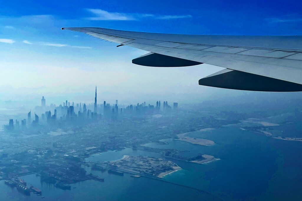 Plane wing seen from a window seat over Dubai, with the port and coast below and the Burj Khalifa in the hazy skyline